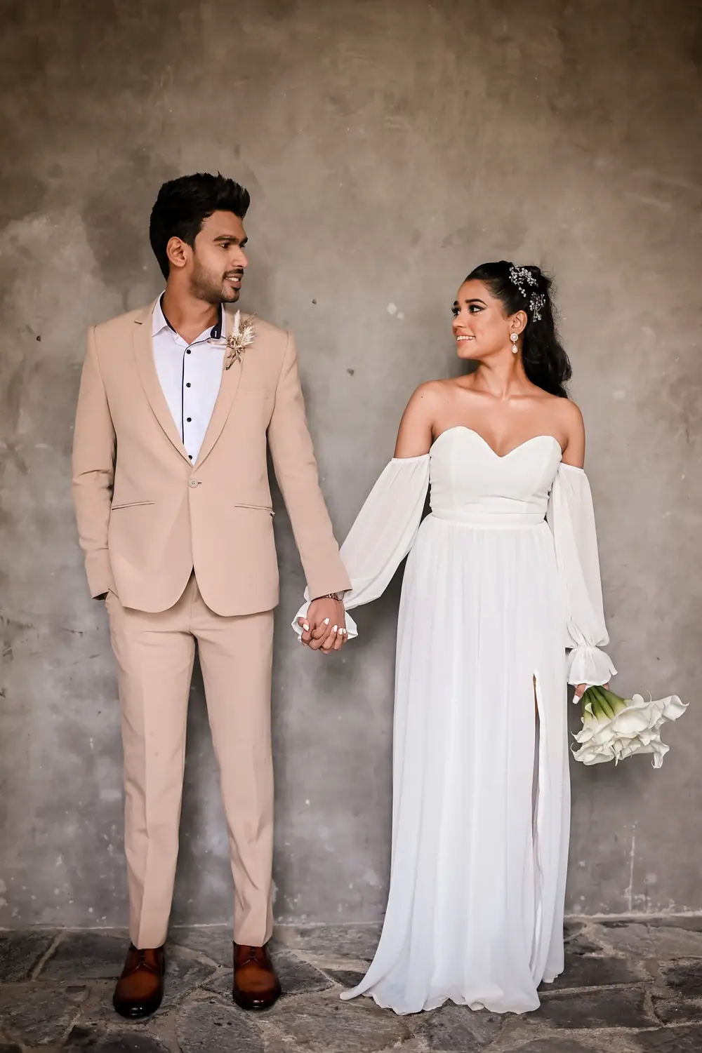 Couple holding hands against a textured concrete wall, he in a beige suit and she in a white dress