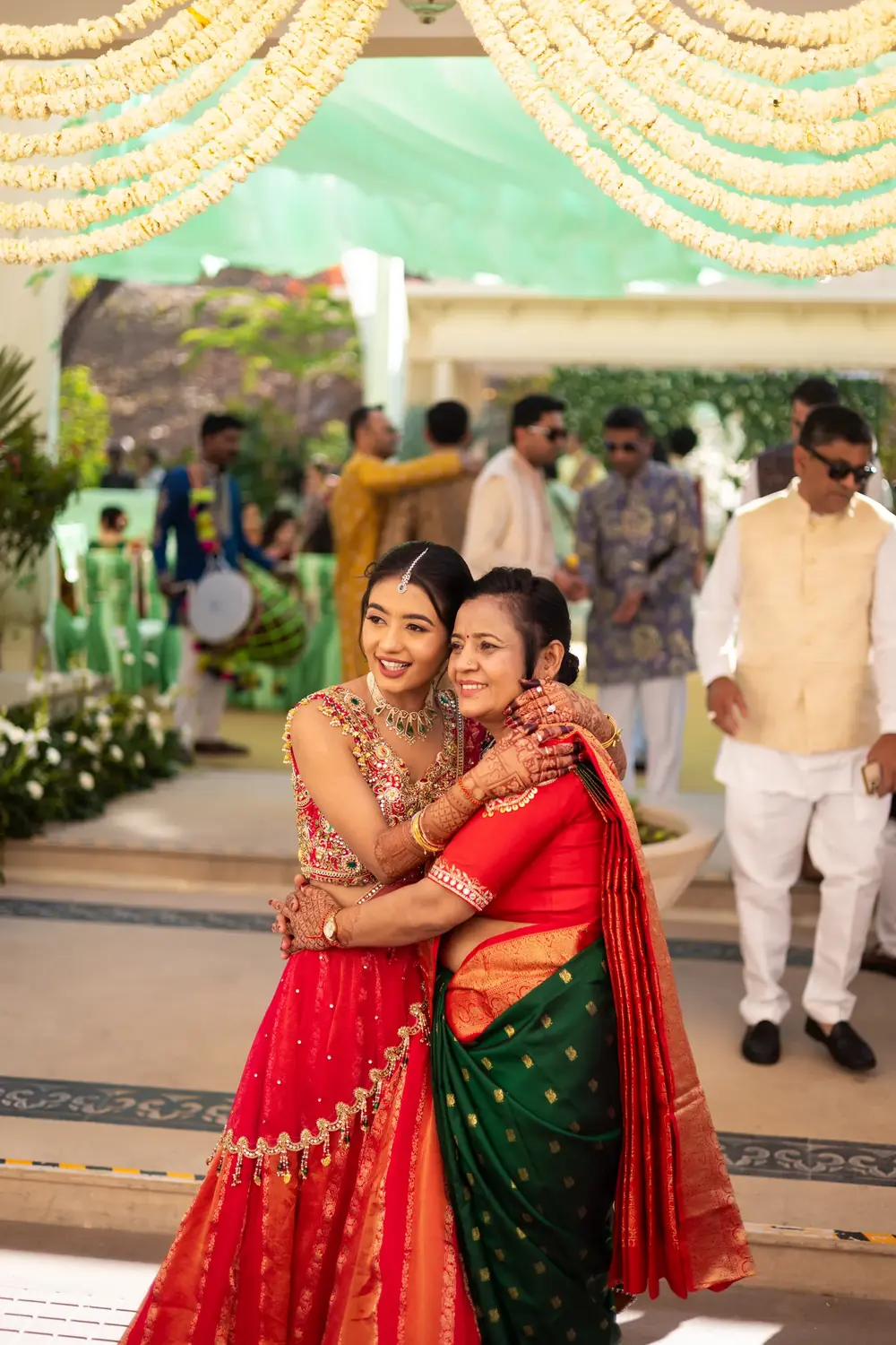 Two women in red and green saris sharing a warm hug beneath a floral mandap