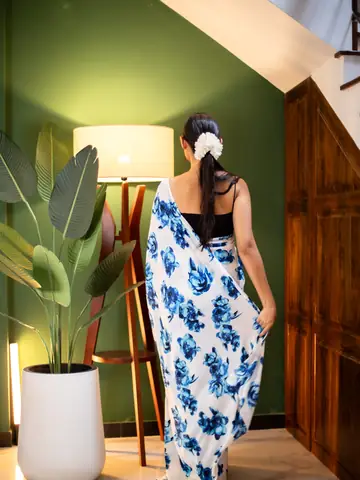 Model in a blue and white floral saree beside a lamp and plant against a green wall