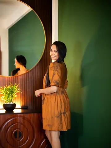 Model in a rust-brown dress beside a round mirror and a green wall