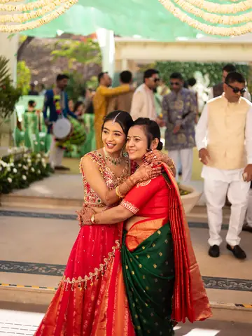Two women in red and green saris sharing a warm hug beneath a floral mandap