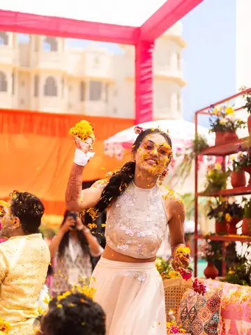 Bride laughing with arms raised as marigold petals fall at the haldi