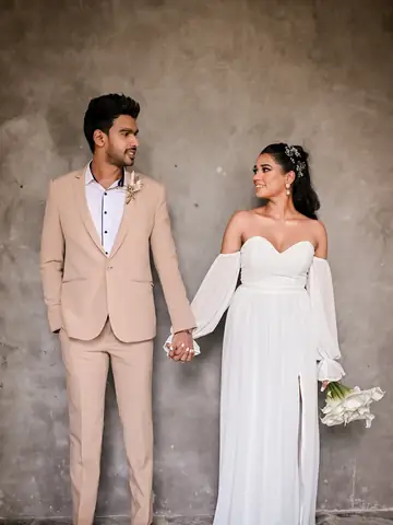 Couple holding hands against a textured concrete wall, he in a beige suit, she in white