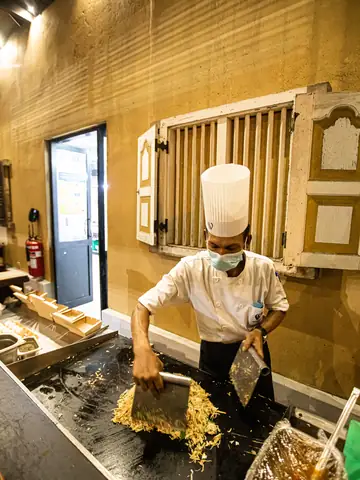 Chef in a tall white toque chopping kottu on a hot griddle at an open kitchen