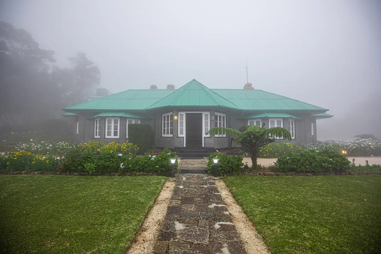 Colonial-style bungalow with a green roof at the end of a stone path across a misty lawn