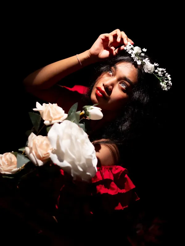 Renaissance-inspired portrait of a model in red lying back on a black backdrop, tucking white flowers into her dark hair